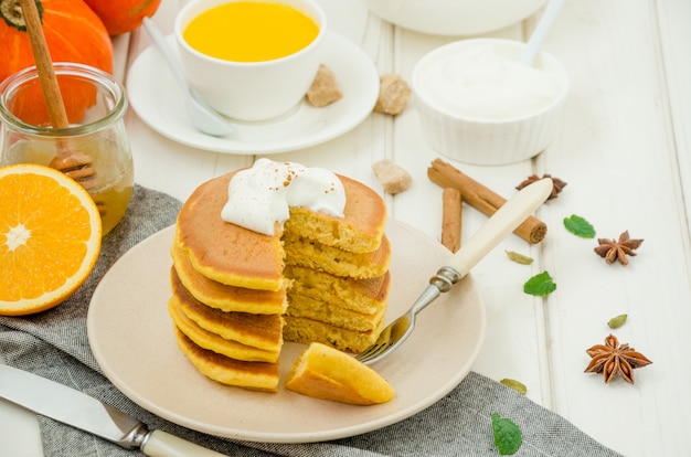 Stapel würzige Kürbispfannkuchen auf einer Platte mit Honig und Schlagsahne auf einer weißen Holzoberfläche