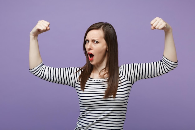 Starke junge Frau in lässig gestreifter Kleidung posiert isoliert auf violettem Hintergrund Studioportrait. People-Lifestyle-Konzept. Mock-up-Kopienbereich. Mund offen halten, Bizeps, Muskeln zeigen.