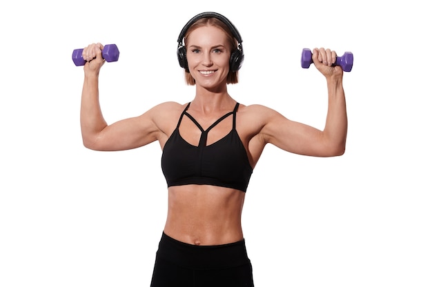 Starke muskulöse Sportlerin in Kopfhörern und schwarzem Outfit, die Übung mit Hanteln auf weißem, isoliertem Hintergrund macht