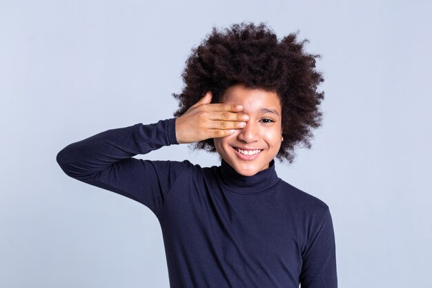 Starke weiße Zähne. Lächelndes, lockiges, positives Kind, das ein Auge mit der Hand schließt, während es an einem Fotoshooting beteiligt ist