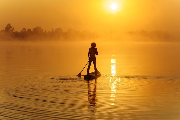 Starker junger Mann in der Badehose, der Paddel verwendet, um während der Morgenzeit auf sup Board zu schwimmen.