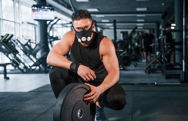 Starker junger Mann in sportlicher Kleidung und gruseliger Maske, der mit schweren Gewichten im Fitnessstudio sitzt