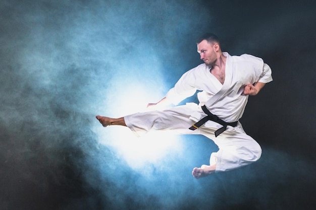 Starker Mann Kämpfer in weißem Kimono für Karate-Kampfkünste mit schwarzem Gürtel Sprung in schwarzem