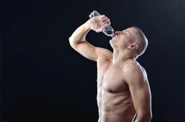 Starker muskulöser Athlet trinkt gierig Wasser aus einer durchsichtigen Plastikflasche auf dunklem Studiohintergrund. Leerer Bereich für Ihren Text.