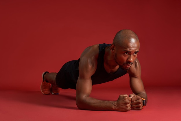 Starker und gutaussehender afrikanischer Mann in Sportkleidung beim Planken isoliert auf rotem Hintergrund