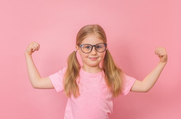 Starkes, erfolgreiches kleines Mädchen mit Brille, das muskulös auf rosa zeigt Gewinner der Gesundheitsstärke