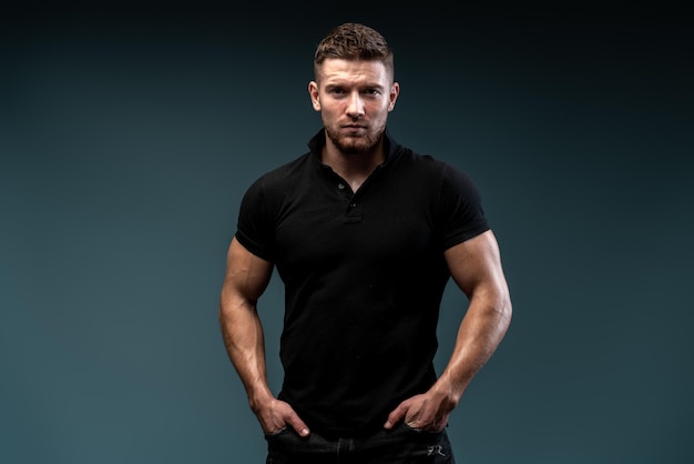 Starkes sportliches Mann-Fitness-Modell mit perfektem Körper, der ein schwarzes T-Shirt trägt, das einzeln auf dunklem Hintergrund posiert. Bodybuilding-Konzept