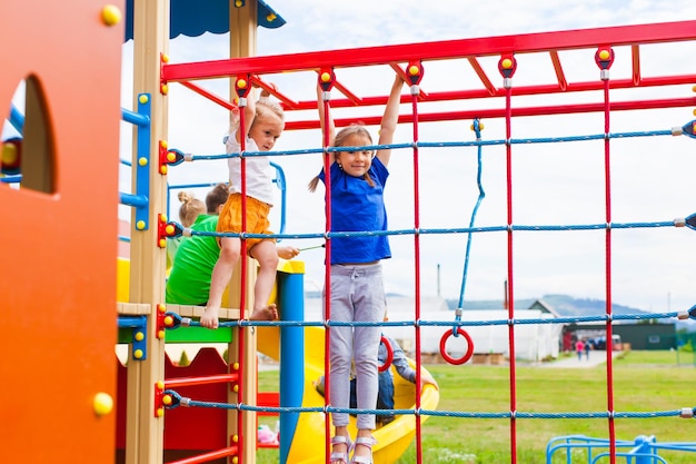 Starkes und gesundes Vorschulmädchen, das an einer horizontalen Kletterleiter aus Metall hängt. Rotes Kletternetz auf einem Vordergrund. Spielplatz im Freien