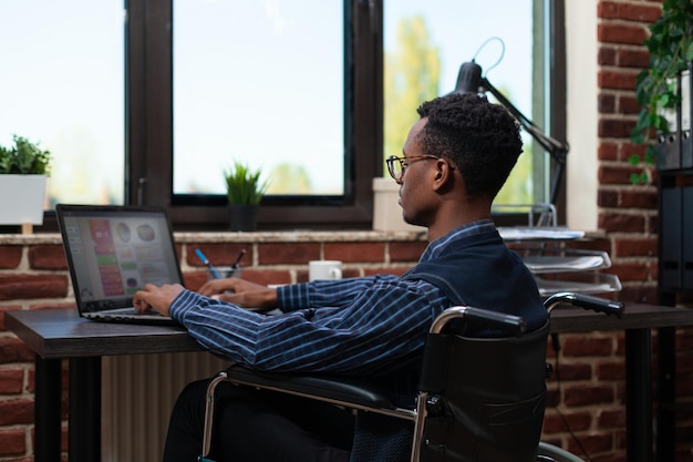 Startup-Angestellter, der im Rollstuhl arbeitet und am Laptop mit Geschäftsanalysen in einem Büro aus rotem Backstein arbeitet. Afroamerikanischer Marketingspezialist, der wichtige Leistungsindikatoren analysiert.