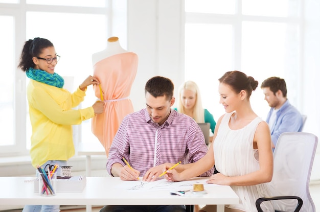 Startup-, Bildungs-, Mode- und Bürokonzept - lächelnde Designer, die Skizzen zeichnen und das Kleid auf der Schaufensterpuppe im Büro anpassen