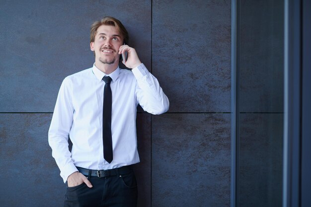 Startup-Geschäftsmann in einem weißen Hemd mit schwarzer Krawatte mit Handy, während er während der Pause von der Arbeit draußen vor einer grauen Wand steht