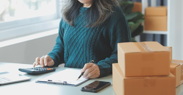 Startup PME pequeno empresário de mulher asiática freelance usando avental usando laptop e caixa para receber e revisar pedidos on-line para se preparar para embalar vender para clientes ideias de negócios on-line para PME