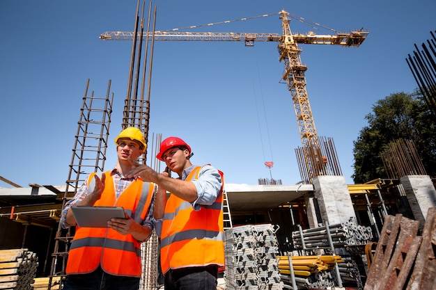 Statiker und Architekt in orangefarbenen Arbeitswesten und harten Schlägern besprechen den Bauablauf per Telefon und Tablet auf der offenen Baustelle in der Nähe des Krans. .