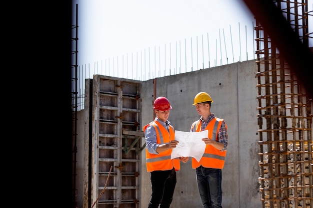 Statiker und Bauleiter in orangefarbener Arbeitsweste und Schutzhelm erkunden die Baudokumentation auf der Baustelle in der Nähe der Stahlrahmen. .