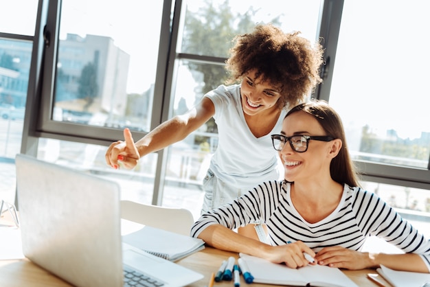 Éste. Hermosas amigas positivas que usan la computadora portátil y se sientan en la mesa mientras estudian juntos