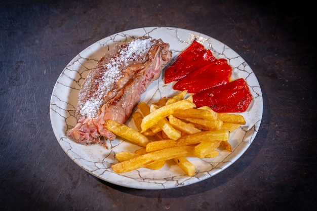 Steak mit Kartoffeln und Paprika auf schwarzem Hintergrund, auf einem weißen Teller