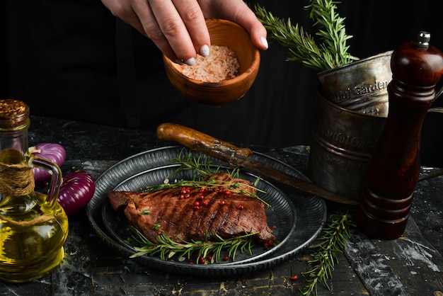 Steak mit Pfeffer und Rosmarin in den Händen des Küchenchefs auf schwarzem Steinhintergrund