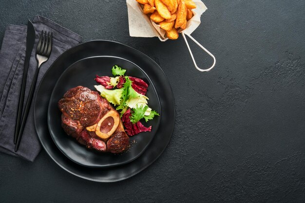 Steaks Osso Buco Geschnittenes Rindfleisch gegrilltes Osso Buco oder Porterhouse-Fleischsteak mit Garnierung mit Salat und Pommes Frites auf schwarzem Marmorbrett auf altem Holzhintergrund Draufsicht Mock up