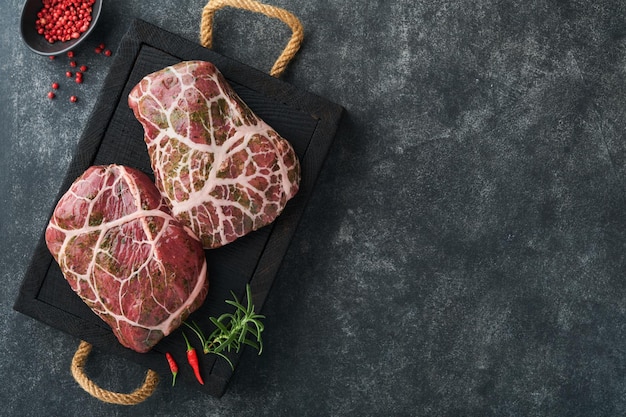 Steaks roh Rohes Cowboysteak marmoriertes Rindfleisch mit Gewürzen Rosmarin und Pfeffer auf schwarzem Holzbrett auf altem schwarzem Schiefertisch Hintergrund Draufsicht Mock up