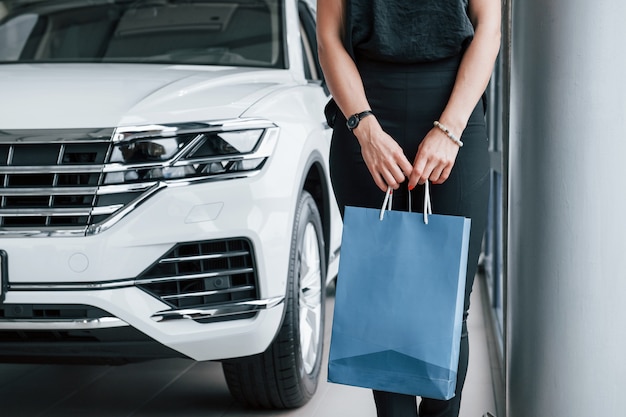 Stehend mit Tasche in den Händen. Mädchen und modernes Auto im Salon. Tagsüber drinnen. Neuwagen kaufen.