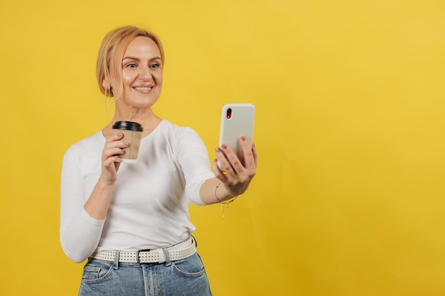Stehendes Lächeln der reifen Frau unter Verwendung des Handys, das sie hält, Kaffee-Pappbecher