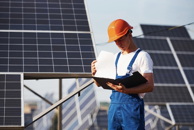 Steht mit Notizblock. Männlicher Arbeiter in blauer Uniform im Freien mit Solarbatterien am sonnigen Tag.