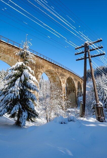 Steinerne Viadukt-Bogenbrücke auf Eisenbahn durch schneebedeckten Tannenwald in den Bergen Schneeverwehungen am Wegesrand und Rauhreif auf Bäumen und elektrischen Leitungen