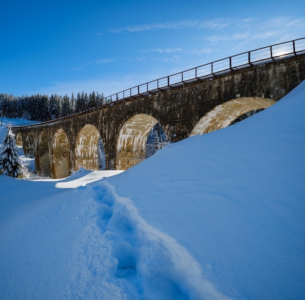 Steinerne Viadukt-Bogenbrücke auf Eisenbahn durch schneebedeckten Tannenwald in den Bergen Schneeverwehungen am Wegesrand und Rauhreif auf Bäumen