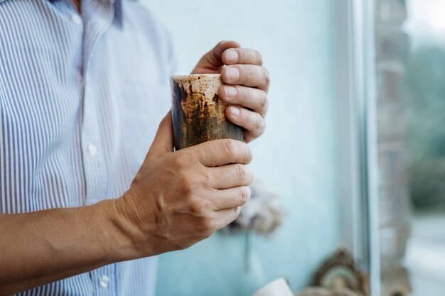 Steingutbecher. Mann, der nahe dem Fenster steht und Tonbecher mit Tee in seinen Händen hält