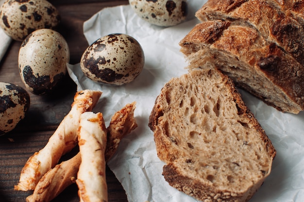 Stellen Sie dunkles hefefreies Buchweizenbrot in einen Schnitt auf Pergament, neben Wachteleiern und italienischen Grissini auf eine Leinentischdecke auf einem Holztisch im rustikalen Stil. Frühstückskochkonzept.