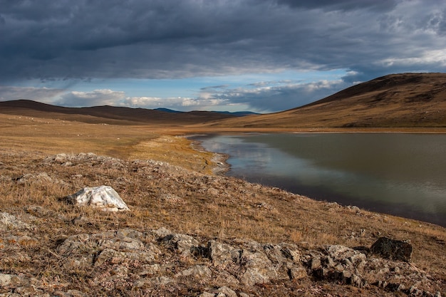 Steppensee mit Steinen im Vordergrund und Hügeln in der Ferne, Wolken am Himmel