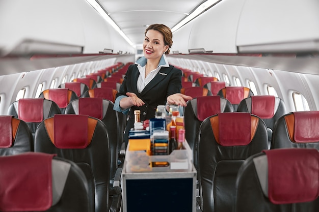 Stewardess, die Essenswagen in der Passagierkabine des Flugzeugjets zeigt. Modernes Flugzeuginterieur. Lächelnde europäische Frau trägt Uniform und schaut in die Kamera. Zivile kommerzielle Luftfahrt. Flugreisekonzept