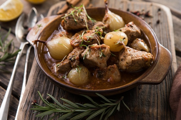 Stifado - köstlicher mediterraner Rindfleischeintopf mit Zwiebelknollen, Zimt und Gewürzen in einer Auflaufform, auf einem schwarzen Holztisch, Ansicht von oben, Nahaufnahme