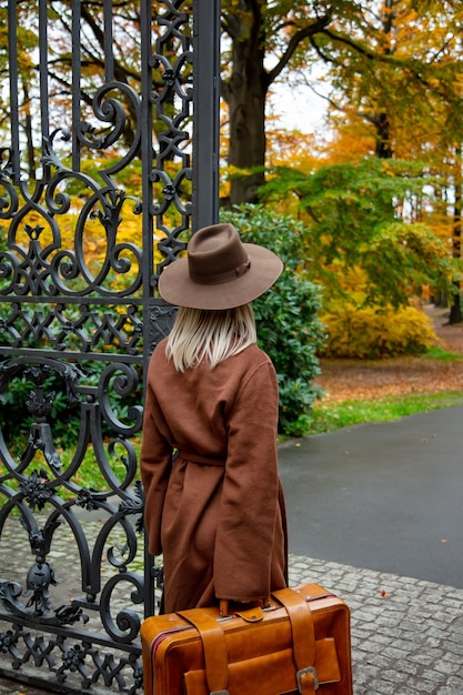 Stil Mädchen im braunen Mantel mit Koffer im Herbstpark nahe Tor