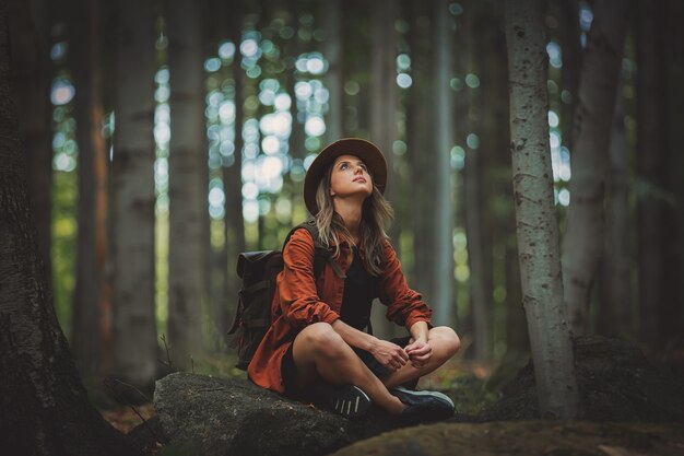 Stil Mädchen mit Rucksack sitzen in einem Stein in einem Sommerzeit gemischten Nadelwald