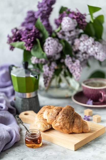 Stillleben-Croissants und eine Tasse Kaffee und ein Strauß Flieder auf dem Tisch am Fenster.