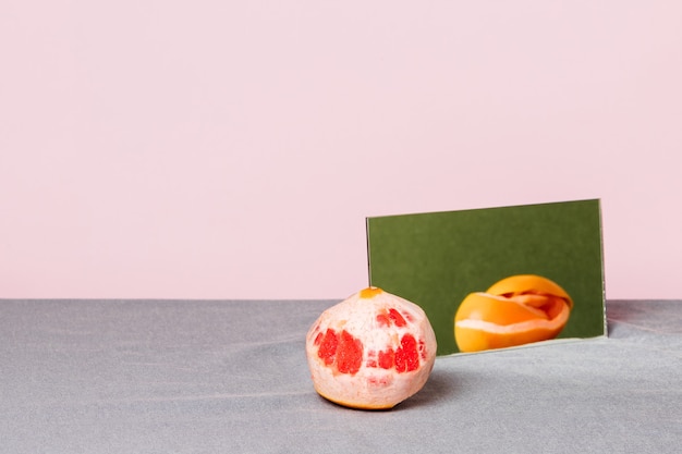 Stillleben einer geschälten Grapefruit, deren Schale sich auf einem Spiegel auf einer grauen Tischdecke und einem Rosa widerspiegelt