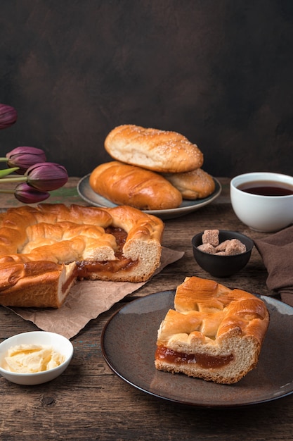 Stillleben mit Gebäck, Kuchen mit Füllung, Brötchen und Tee auf braunem Grund. Seitenansicht, vertikal