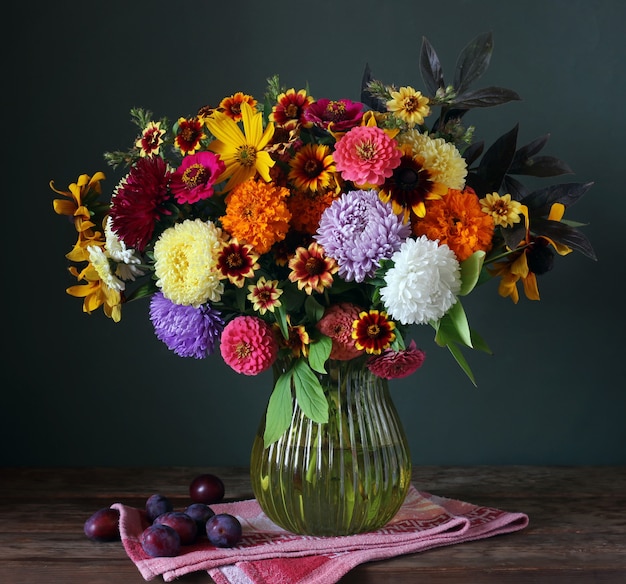 Stillleben mit Herbstblumenstrauß in einer Vase und Pflaumen auf Dunkelheit