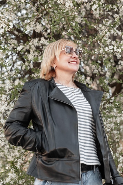 Stilvoll schöne und glückliche ältere Frau mit einem Lächeln in modischer Freizeitkleidung mit schwarzer Lederjacke und T-Shirt, die in der Nähe eines blühenden Baums im Park spaziert