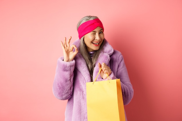 Stilvolle ältere asiatische Frau, die Geschäft empfiehlt, Einkaufstasche hält und mit OK-Zeichen zwinkert, zufrieden lächelt und auf rosa Hintergrund steht