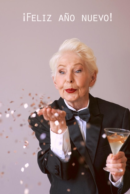 Stilvolle ältere Frau im Smoking mit einem Glas Champagner, das Konfetti bläst und das neue Jahr feiert