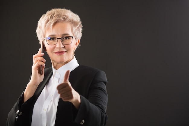 stilvolle ältere Frau in Brille und in einer schwarzen Jacke ruft mit einer Handbewegung am Telefon an