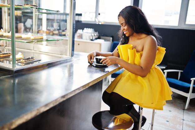 Stilvolle afrikanisch-amerikanische Frau im gelben Kleid posierte im Café mit einer Tasse Cappuccino