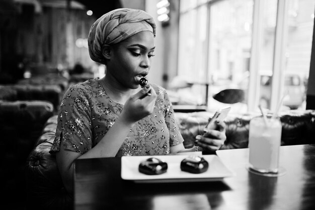 Stilvolle afrikanische Frau in rotem Hemd und Hut posierte im Innencafé, isst Schokoladendessertkuchen und schaut aufs Handy