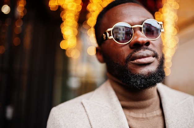 Stilvolle Afroamerikaner tragen beige Jacke mit Sonnenbrille gegen Haus mit goldener Girlande.