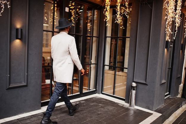 Stilvolle Afroamerikaner tragen beige Jacke mit Sonnenbrille gegen Haus mit goldener Girlande.