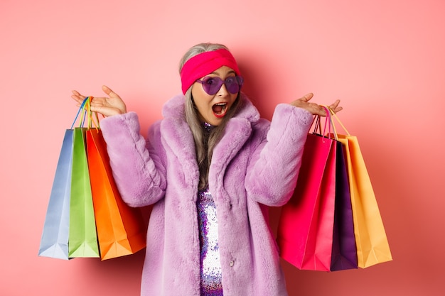Stilvolle asiatische Seniorin, die einkaufen geht, trendige Kleidung und Sonnenbrille trägt, Ladentaschen mit Geschenken hält, sich selbst ein Konzept gönnt, rosa Hintergrund