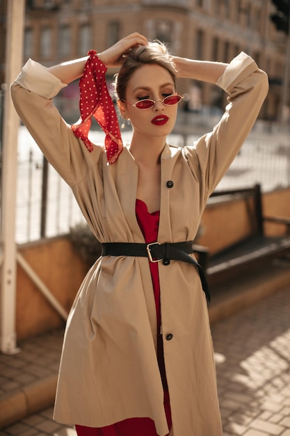 Stilvolle attraktive blonde Frau mit roten Lippen berührt Haare Elegante Dame in hellem Sonnenbrillen-Seidenkleid und beigem Trenchcoat posiert draußen