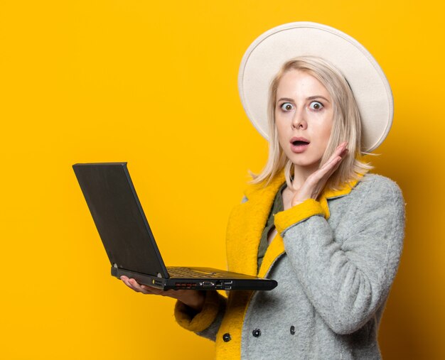 Stilvolle blonde Frau im Hut und im saisonalen Mantel mit Laptop